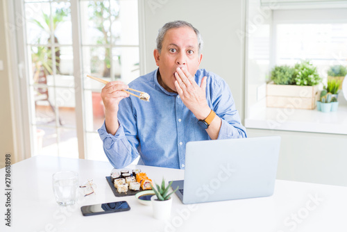 Handsome business senior man eating delivery sushi while working using laptop cover mouth with hand shocked with shame for mistake, expression of fear, scared in silence, secret concept