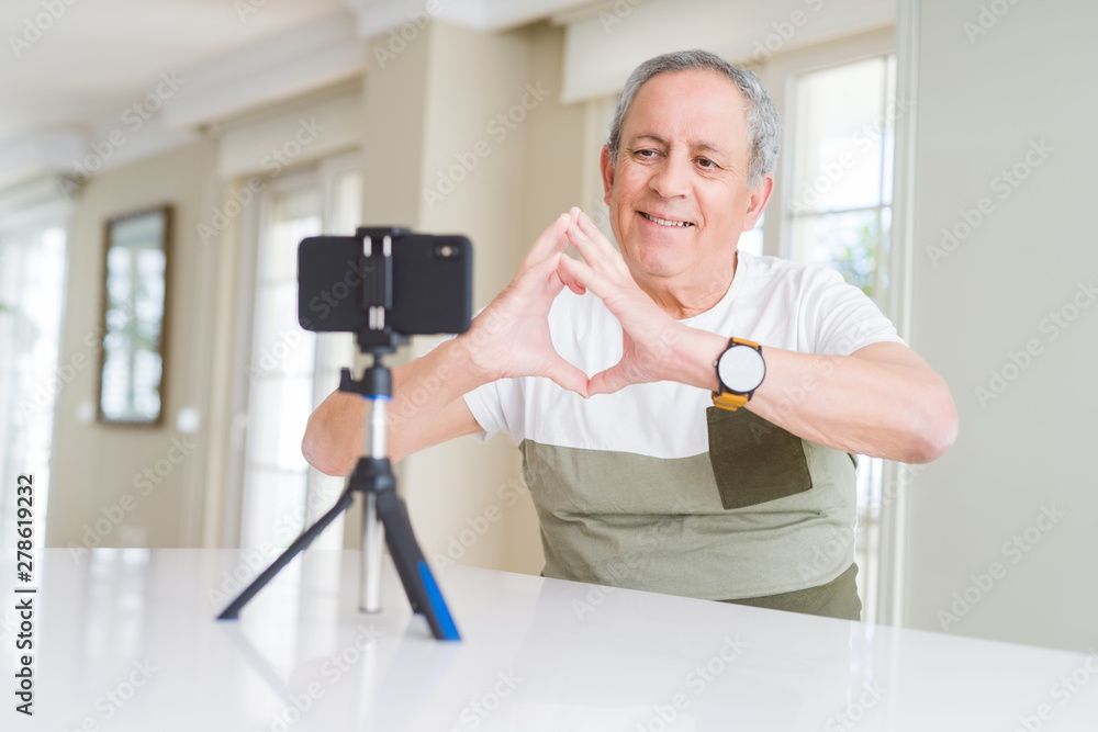 Handsome senior man doing video conference talking to the smartphone camera smiling in love showing heart symbol and shape with hands. Romantic concept. Handsome senior man doing video conference talking to the smartphone camera smiling in love showing heart symbol and shape with hands. Romantic concept.