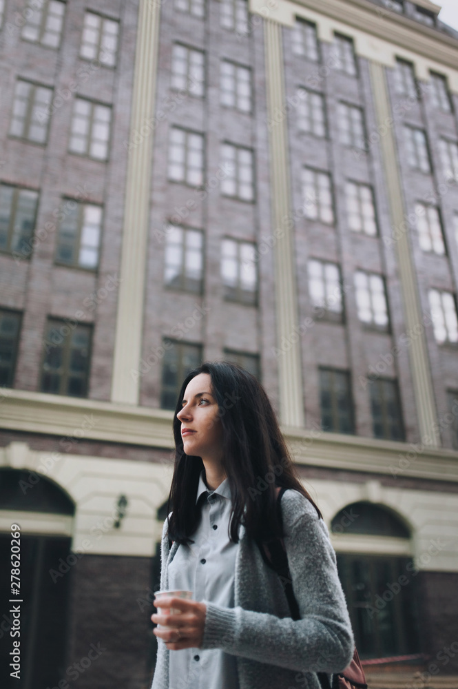 Young beautiful caucasian girl walking along the street with backpack and paper cup of coffee. The concept of loneliness, sadness and unemployment. Vertical, bottom view. Young beautiful caucasian girl walking along the street with backpack and paper cup of coffee. The concept of loneliness, sadness and unemployment. Vertical, bottom view.