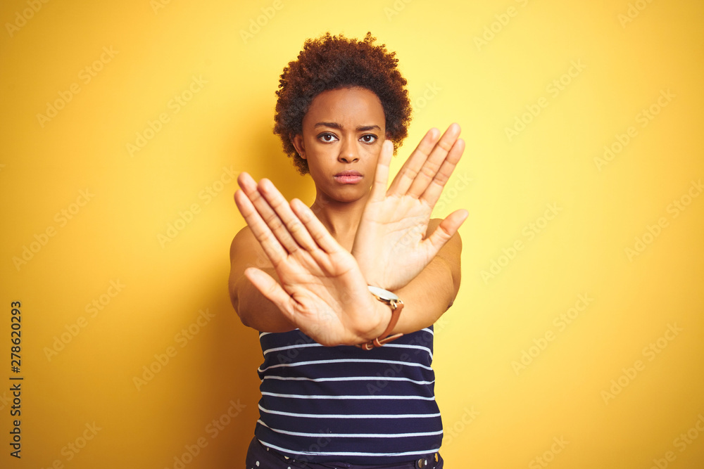Beauitul african american woman wearing summer t-shirt over isolated yellow background Rejection expression crossing arms and palms doing negative sign, angry face Beauitul african american woman wearing summer t-shirt over isolated yellow background Rejection expression crossing arms and palms doing negative sign, angry face