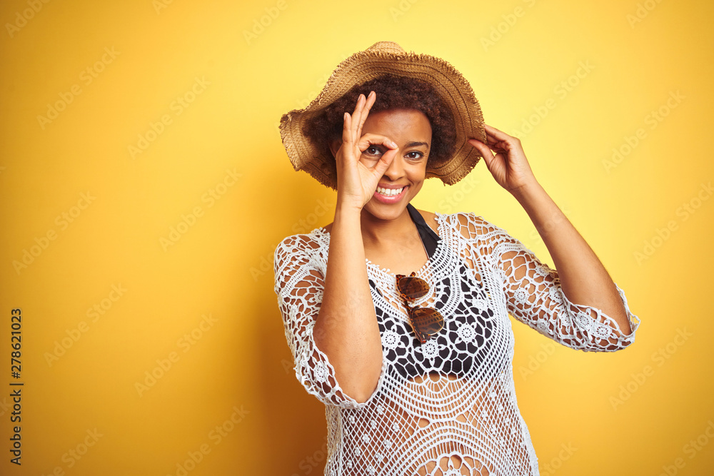Young african american woman with afro hair wearing summer hat over white isolated background doing ok gesture with hand smiling, eye looking through fingers with happy face. Young african american woman with afro hair wearing summer hat over white isolated background doing ok gesture with hand smiling, eye looking through fingers with happy face.