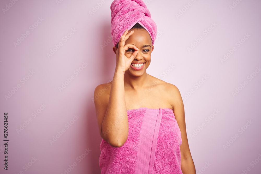 African american woman wearing shower towel after bath over pink isolated background doing ok gesture with hand smiling, eye looking through fingers with happy face. African american woman wearing shower towel after bath over pink isolated background doing ok gesture with hand smiling, eye looking through fingers with happy face.