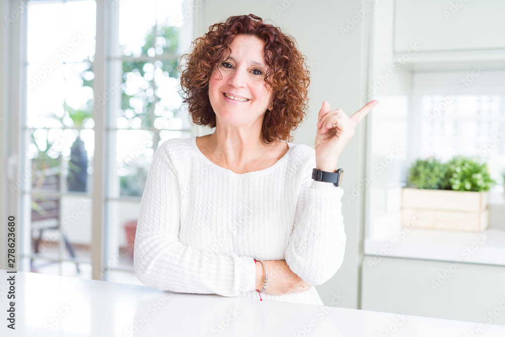 Beautiful senior woman wearing white sweater at home with a big smile on face, pointing with hand and finger to the side looking at the camera. Beautiful senior woman wearing white sweater at home with a big smile on face, pointing with hand and finger to the side looking at the camera.