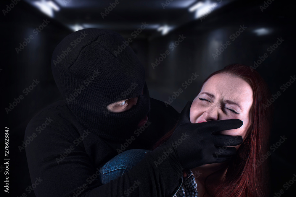 Scared young victim woman caught by gangster criminal in dark underground tunnel. Bandit in black mask and gloves covering young woman's mouth with his hand. Kidnapping, criminal, terrorist concept. - obrazy, fototapety, plakaty 