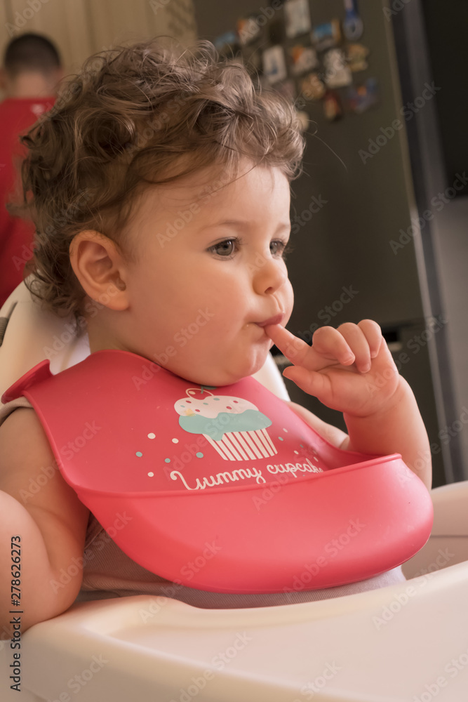 Beautiful baby girl having fun on a feeding chair with a bib on and her father at the background