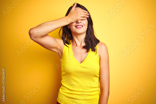 Young beautiful woman wearing t-shirt standing over yellow isolated background smiling and laughing with hand on face covering eyes for surprise. Blind concept.