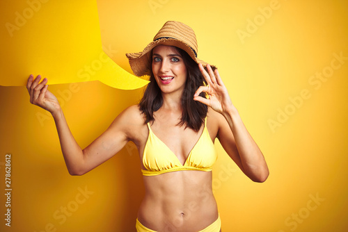 Beautiful woman wearing yellow bikini and holding talking balloon over isolated yellow background doing ok sign with fingers, excellent symbol © Krakenimages.com