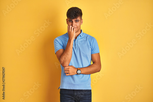 Young indian man wearing blue polo standing over isolated yellow background looking stressed and nervous with hands on mouth biting nails. Anxiety problem.