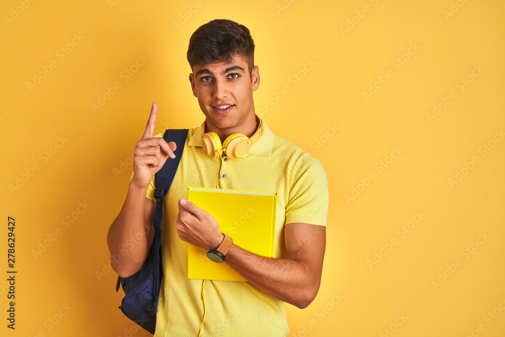 Indian student man wearing backpack headphones notebook over isolated yellow background surprised with an idea or question pointing finger with happy face, number one Indian student man wearing backpack headphones notebook over isolated yellow background surprised with an idea or question pointing finger with happy face, number one