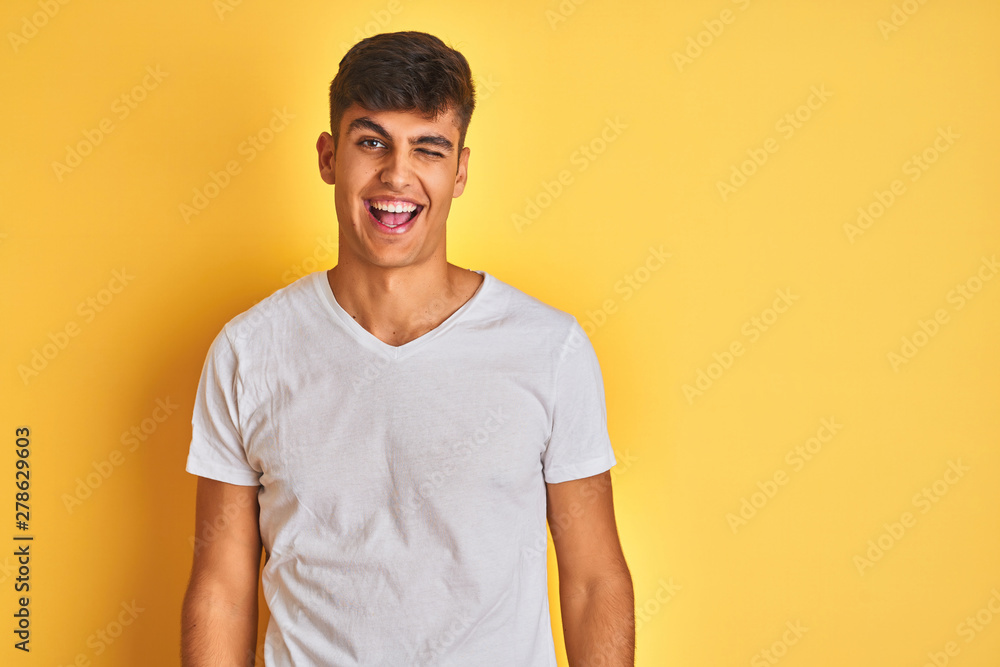 Young indian man wearing white t-shirt standing over isolated yellow background winking looking at the camera with sexy expression, cheerful and happy face. Young indian man wearing white t-shirt standing over isolated yellow background winking looking at the camera with sexy expression, cheerful and happy face.
