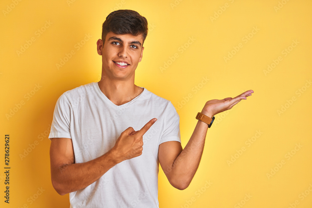 Young indian man wearing white t-shirt standing over isolated yellow background amazed and smiling to the camera while presenting with hand and pointing with finger. Young indian man wearing white t-shirt standing over isolated yellow background amazed and smiling to the camera while presenting with hand and pointing with finger.