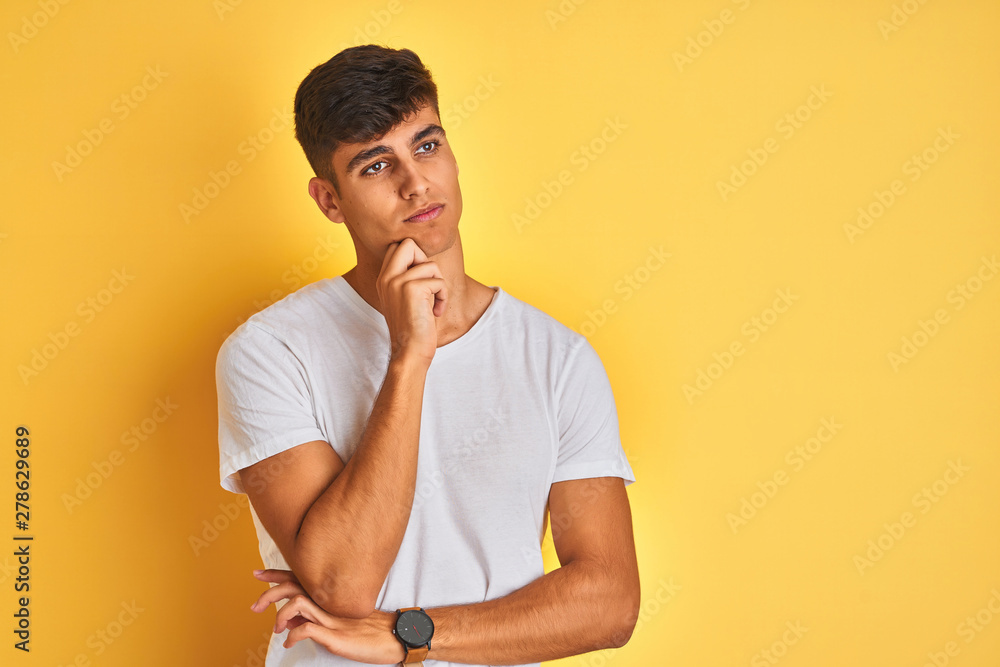Young indian man wearing white t-shirt standing over isolated yellow background with hand on chin thinking about question, pensive expression. Smiling with thoughtful face. Doubt concept. Young indian man wearing white t-shirt standing over isolated yellow background with hand on chin thinking about question, pensive expression. Smiling with thoughtful face. Doubt concept.