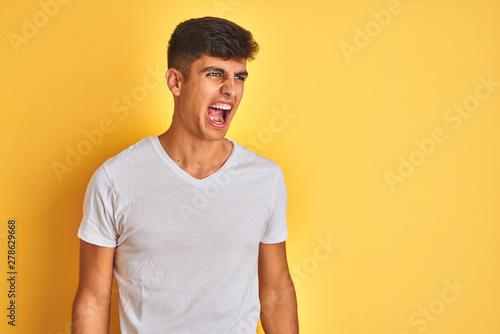 Young indian man wearing white t-shirt standing over isolated yellow background angry and mad screaming frustrated and furious, shouting with anger. Rage and aggressive concept.
