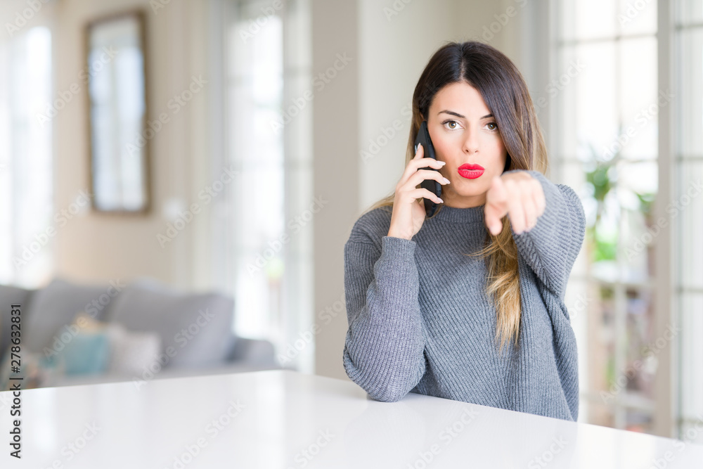 Young beautiful woman talking on the phone at home pointing with finger to the camera and to you, hand sign, positive and confident gesture from the front Young beautiful woman talking on the phone at home pointing with finger to the camera and to you, hand sign, positive and confident gesture from the front