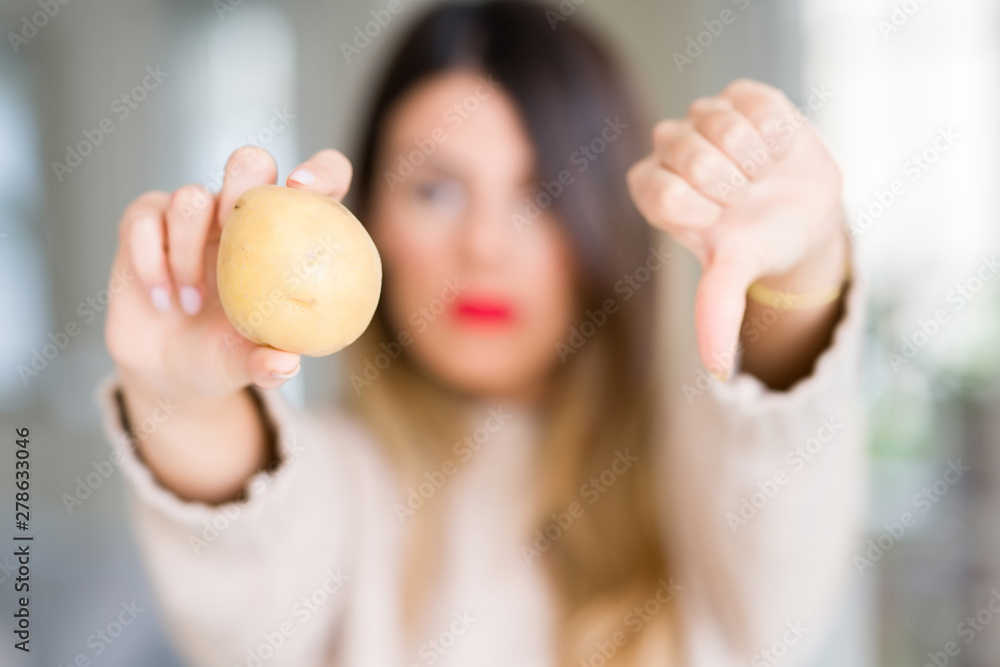 Young beautiful woman holding fresh potato at home with angry face, negative sign showing dislike with thumbs down, rejection concept Young beautiful woman holding fresh potato at home with angry face, negative sign showing dislike with thumbs down, rejection concept