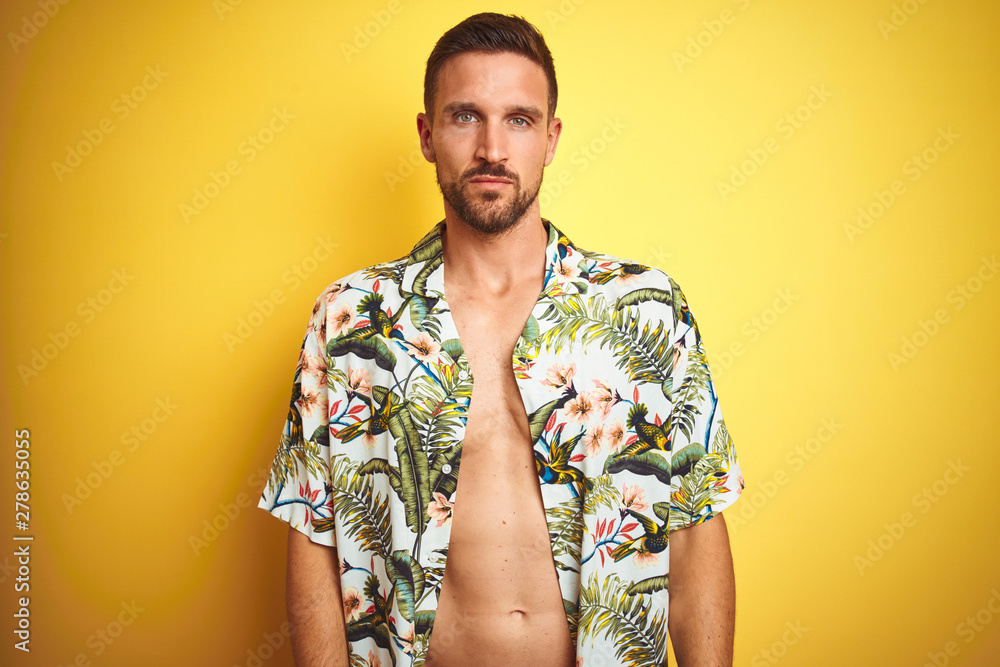 Handsome man wearing summer hawaiian flowers shirt over yellow isolated background with serious expression on face. Simple and natural looking at the camera. Handsome man wearing summer hawaiian flowers shirt over yellow isolated background with serious expression on face. Simple and natural looking at the camera.