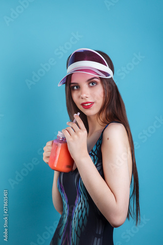 Sexy woman in swimsuit and plastic visor holding glass of fresh summer drink on blue background