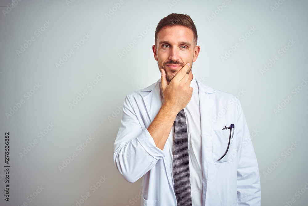 Young handsome doctor man wearing white profressional coat over isolated background looking confident at the camera with smile with crossed arms and hand raised on chin. Thinking positive. Young handsome doctor man wearing white profressional coat over isolated background looking confident at the camera with smile with crossed arms and hand raised on chin. Thinking positive.