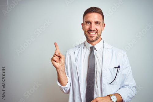 Young handsome doctor man wearing white profressional coat over isolated background with a big smile on face, pointing with hand and finger to the side looking at the camera. photo