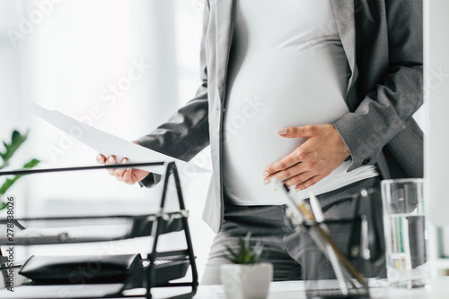 cropped view of pregnant woman holding paper and belly, standing near table with document tray and glass
