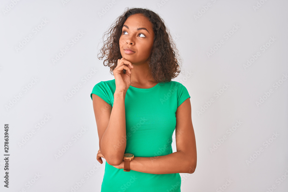Young brazilian woman wearing green t-shirt standing over isolated white background with hand on chin thinking about question, pensive expression. Smiling with thoughtful face. Doubt concept. Young brazilian woman wearing green t-shirt standing over isolated white background with hand on chin thinking about question, pensive expression. Smiling with thoughtful face. Doubt concept.