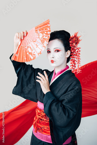 beautiful geisha in black kimono posing with hand fan and red cloth on background isolated on white