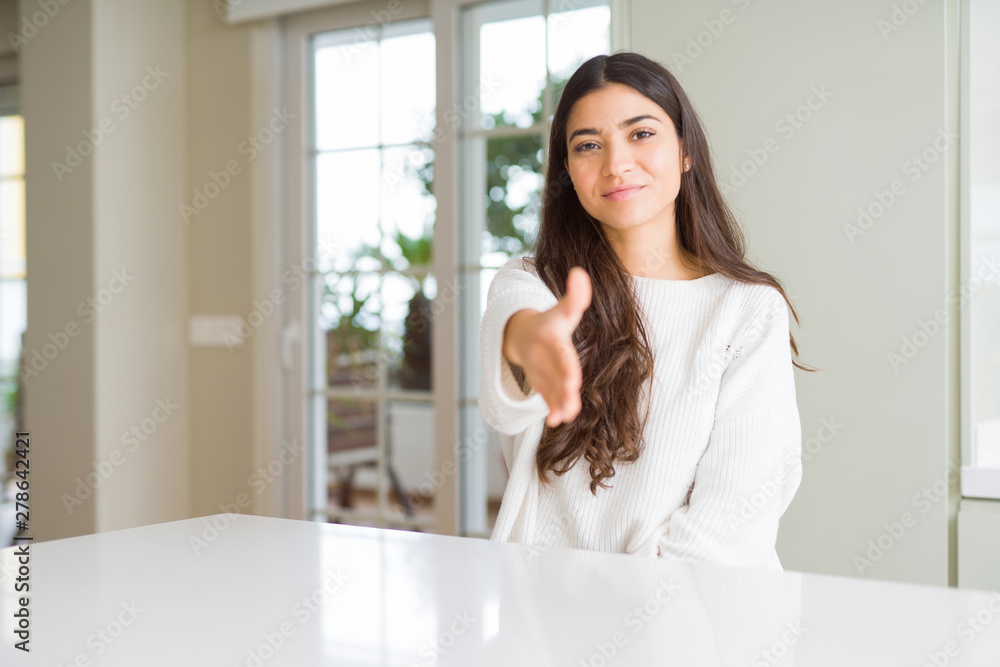 Young beautiful woman at home on white table smiling friendly offering handshake as greeting and welcoming. Successful business.
