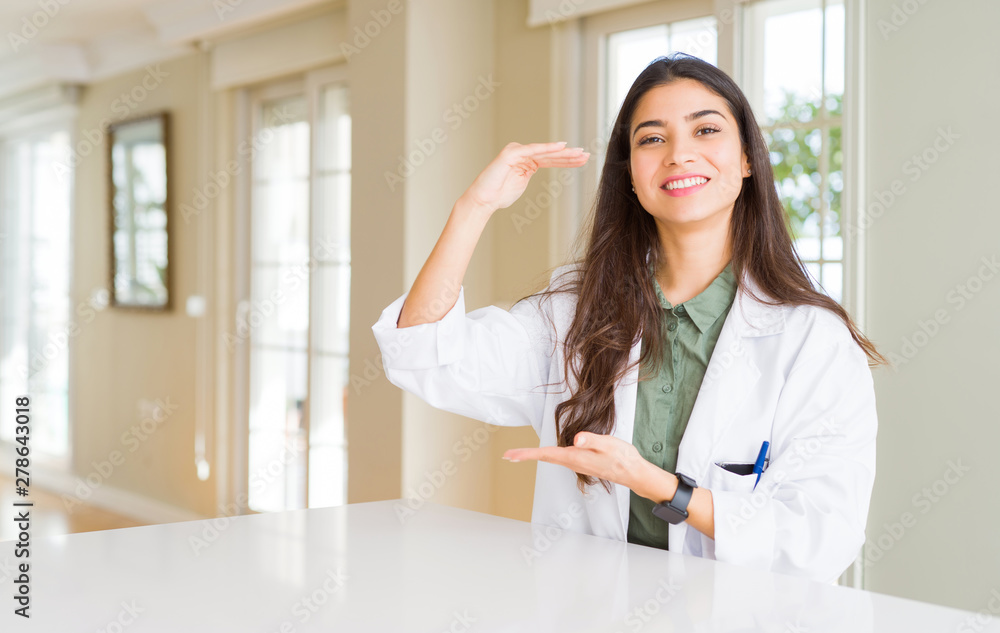 Young woman wearing medical coat at the clinic as therapist or doctor gesturing with hands showing big and large size sign, measure symbol. Smiling looking at the camera. Measuring concept. Young woman wearing medical coat at the clinic as therapist or doctor gesturing with hands showing big and large size sign, measure symbol. Smiling looking at the camera. Measuring concept.