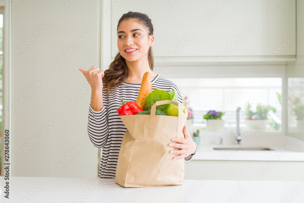 Young woman holding paper bag full of groceries pointing and showing with thumb up to the side with happy face smiling Young woman holding paper bag full of groceries pointing and showing with thumb up to the side with happy face smiling