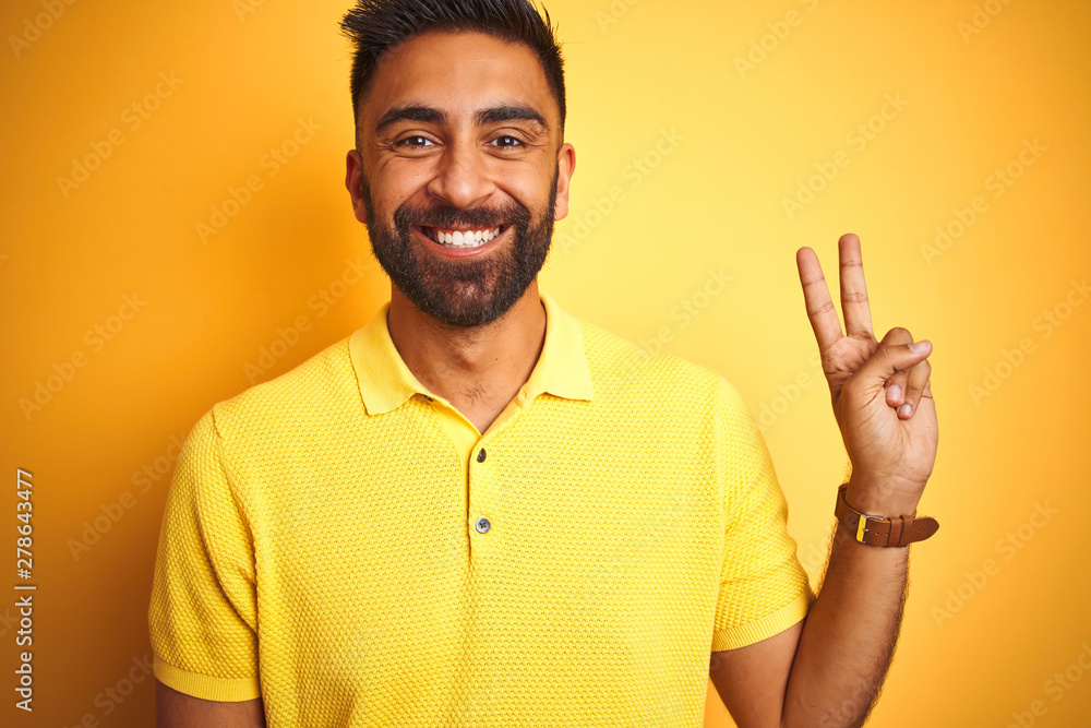 Young indian man wearing polo standing over isolated yellow background smiling with happy face winking at the camera doing victory sign. Number two. Young indian man wearing polo standing over isolated yellow background smiling with happy face winking at the camera doing victory sign. Number two.