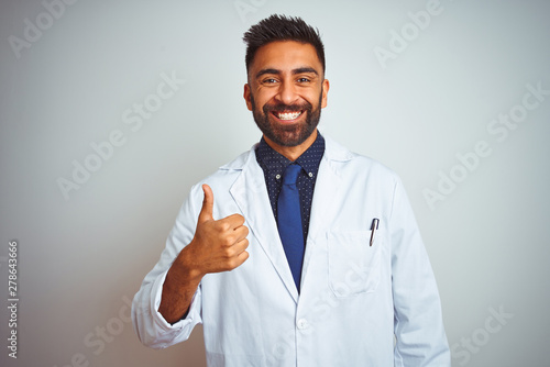 Young indian doctor man standing over isolated white background doing happy thumbs up gesture with hand. Approving expression looking at the camera with showing success.