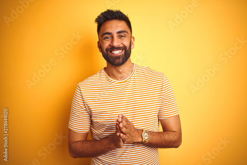 Young indian man wearing t-shirt standing over isolated yellow background Hands together and fingers crossed smiling relaxed and cheerful. Success and optimistic