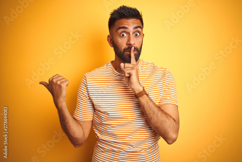 Young indian man wearing t-shirt standing over isolated yellow background asking to be quiet with finger on lips pointing with hand to the side. Silence and secret concept. photo