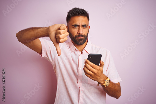 Young indian man using smartphone standing over isolated pink background with angry face, negative sign showing dislike with thumbs down, rejection concept