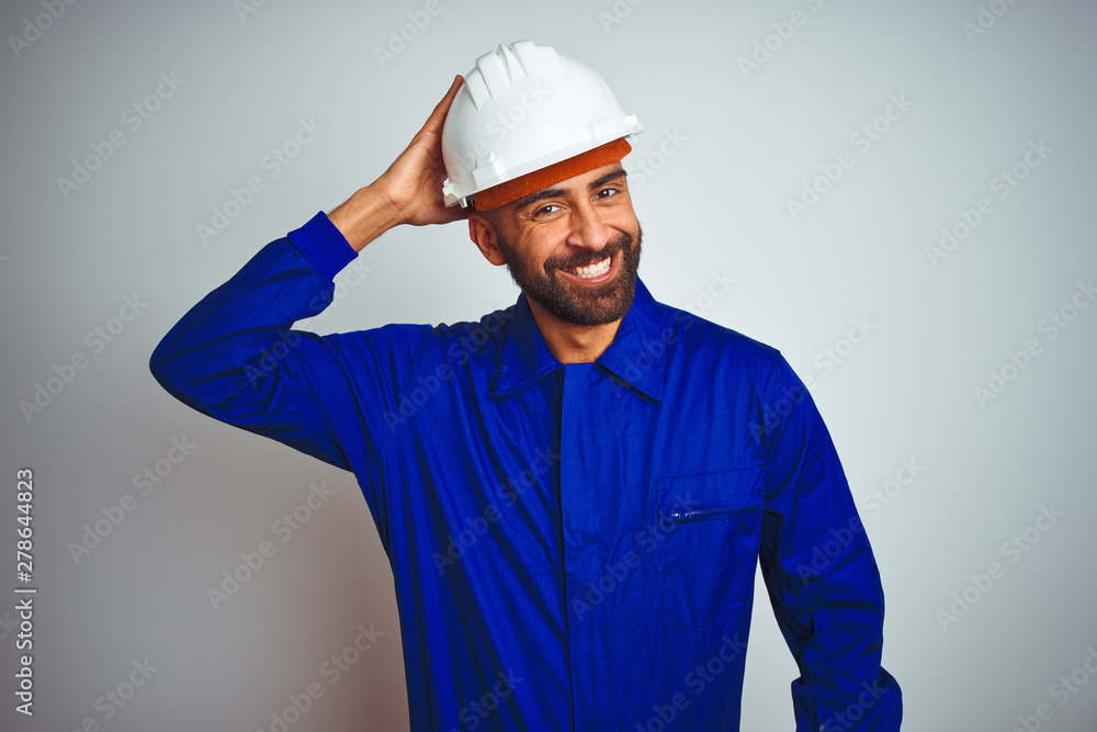 Handsome indian worker man wearing uniform and helmet over isolated white background confuse and wonder about question. Uncertain with doubt, thinking with hand on head. Pensive concept.