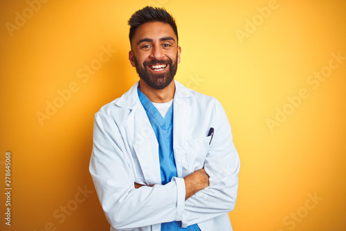 Young indian doctor man standing over isolated yellow background happy face smiling with crossed arms looking at the camera. Positive person.