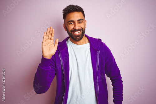 Young indian man wearing purple sweatshirt standing over isolated pink background Waiving saying hello happy and smiling, friendly welcome gesture