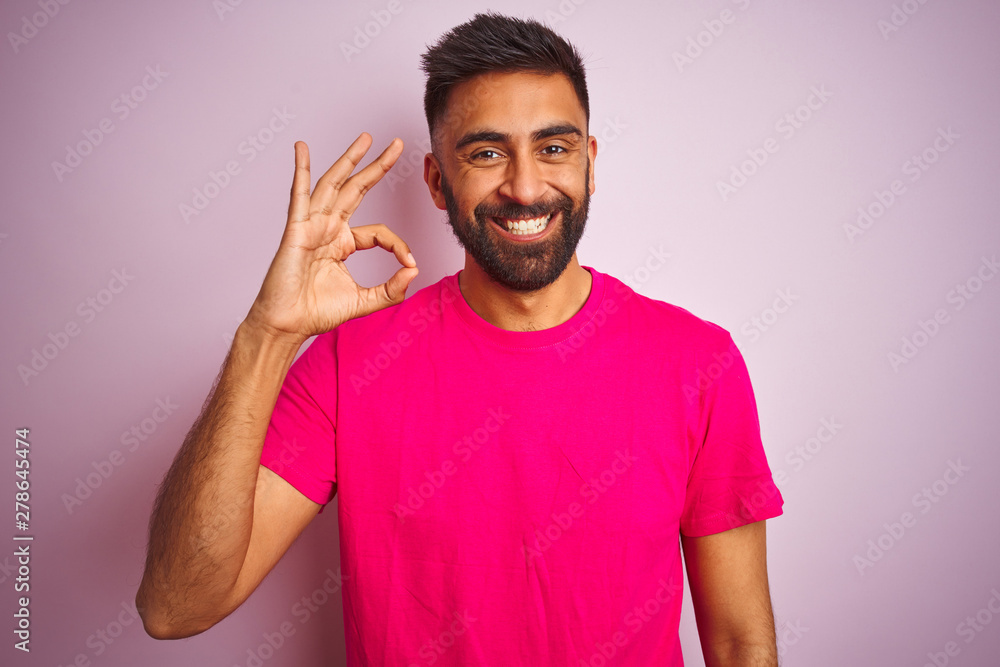 Young indian man wearing t-shirt standing over isolated pink background smiling positive doing ok sign with hand and fingers. Successful expression. Young indian man wearing t-shirt standing over isolated pink background smiling positive doing ok sign with hand and fingers. Successful expression.