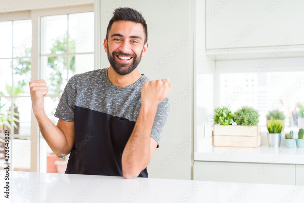 Handsome hispanic man wearing casual t-shirt at home celebrating surprised and amazed for success with arms raised and open eyes. Winner concept.