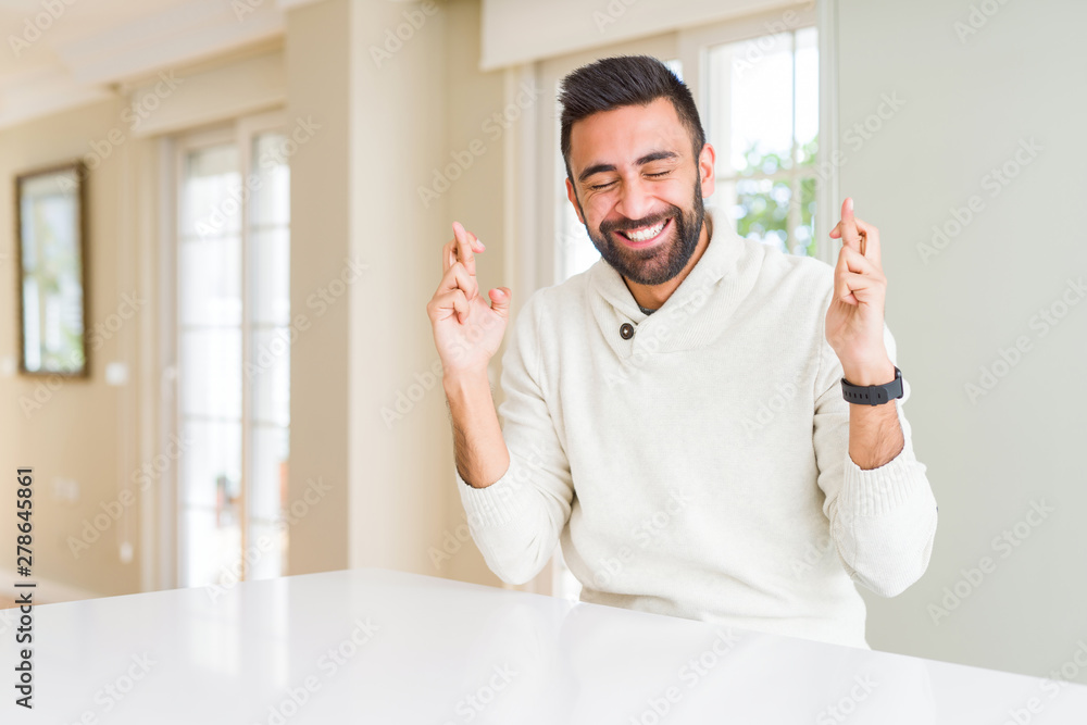 Handsome hispanic man wearing casual white sweater at home smiling crossing fingers with hope and eyes closed. Luck and superstitious concept. Handsome hispanic man wearing casual white sweater at home smiling crossing fingers with hope and eyes closed. Luck and superstitious concept.