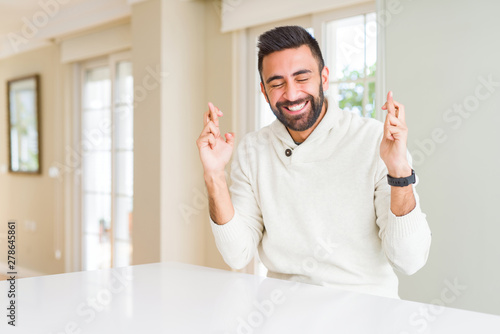 Handsome hispanic man wearing casual white sweater at home smiling crossing fingers with hope and eyes closed. Luck and superstitious concept.