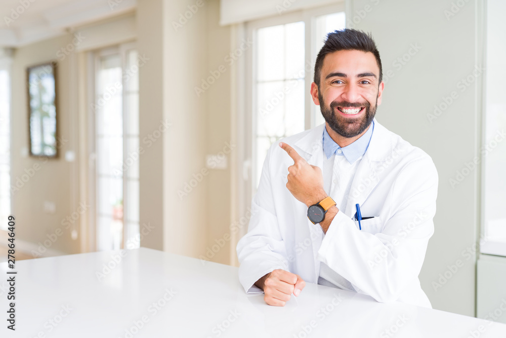 Handsome hispanic doctor or therapist man wearing medical coat at the clinic cheerful with a smile of face pointing with hand and finger up to the side with happy and natural expression on face