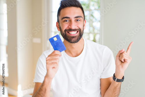 Handsome hispanic man holding credit card very happy pointing with hand and finger to the side