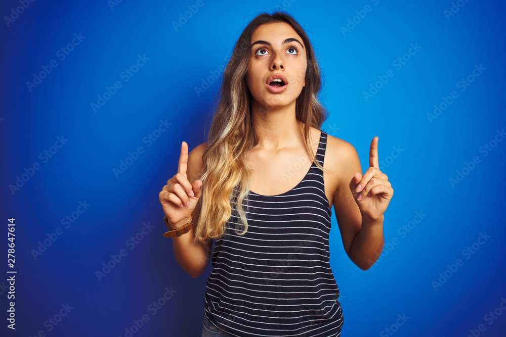 Young beautiful woman wearing stripes t-shirt over blue isolated background amazed and surprised looking up and pointing with fingers and raised arms. Young beautiful woman wearing stripes t-shirt over blue isolated background amazed and surprised looking up and pointing with fingers and raised arms.