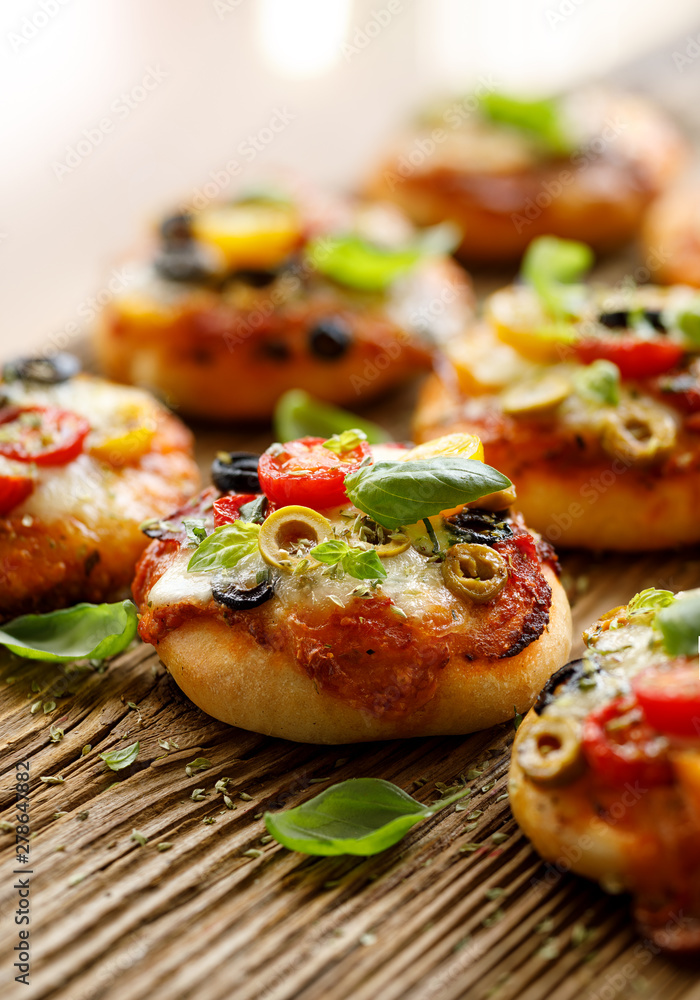 Small pizzas with the addition of cherry tomatoes, olives, mozzarella cheese and fresh basil on a rustic wooden background, close-up. Small pizzas with the addition of cherry tomatoes, olives, mozzarella cheese and fresh basil on a rustic wooden background, close-up.