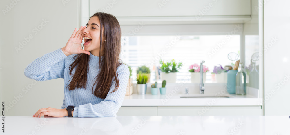 Wide angle picture of beautiful young woman sitting on white table at home shouting and screaming loud to side with hand on mouth. Communication concept. Wide angle picture of beautiful young woman sitting on white table at home shouting and screaming loud to side with hand on mouth. Communication concept.