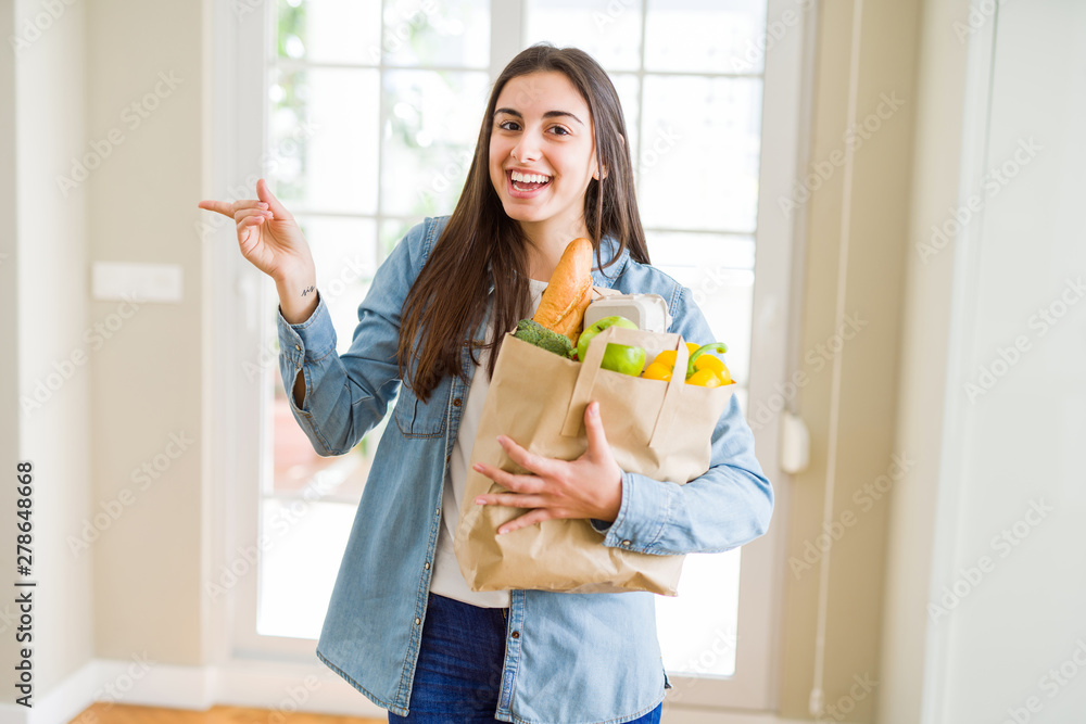 Beautiful young woman holding paper bag full of healthy groceries very happy pointing with hand and finger to the side Beautiful young woman holding paper bag full of healthy groceries very happy pointing with hand and finger to the side