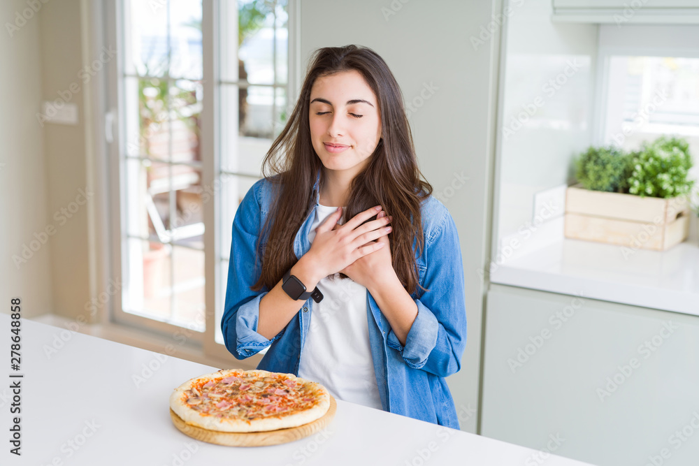 Beautiful young woman eating homemade tasty pizza at the kitchen smiling with hands on chest with closed eyes and grateful gesture on face. Health concept.