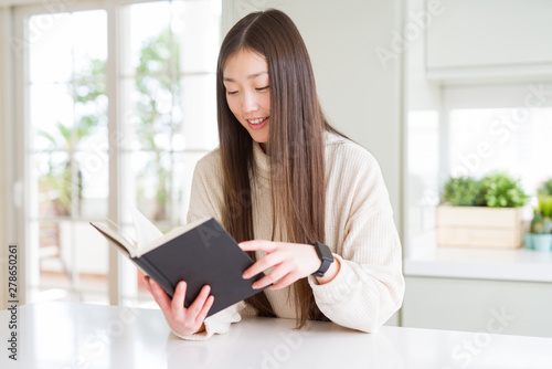 Beautiful Asian woman reading a book with a happy face standing and smiling with a confident smile showing teeth