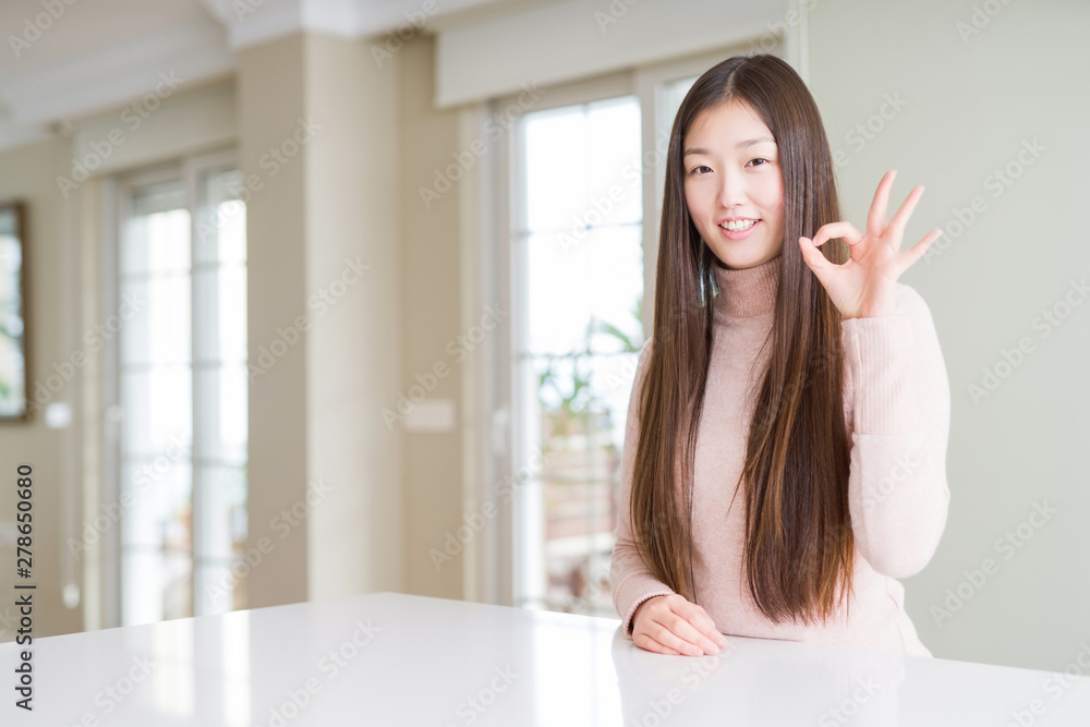 Beautiful Asian woman wearing casual sweater on white table smiling positive doing ok sign with hand and fingers. Successful expression. Beautiful Asian woman wearing casual sweater on white table smiling positive doing ok sign with hand and fingers. Successful expression.